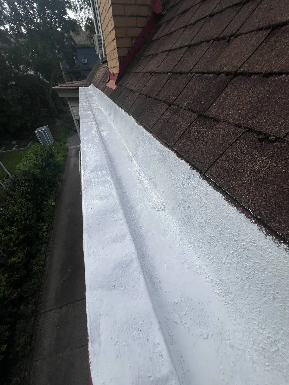 Long view of a gutter with a fresh white sealing coating alongside a shingled roof.